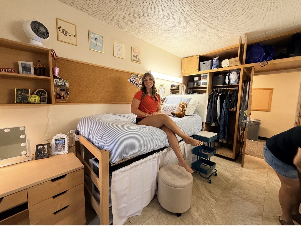 Reese sitting on her made bed in her college dorm room, photos and softball keepsakes on the shelves behind her